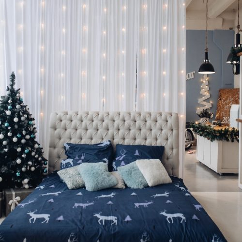 View over made bed with beautiful bedsheet with deer and cushions. The wall decorated with garland. The bedroom decorated for Christmas with fir tree.